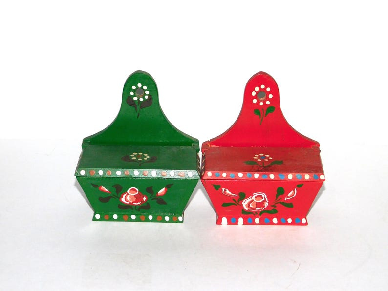May include: Two small wooden wall mounted boxes with painted floral designs. One box is green with white and red flowers and the other is red with white and green flowers. May include: Two small wooden wall mounted boxes with painted floral designs. One box is green with white and red flowers and the other is red with white and green flowers.