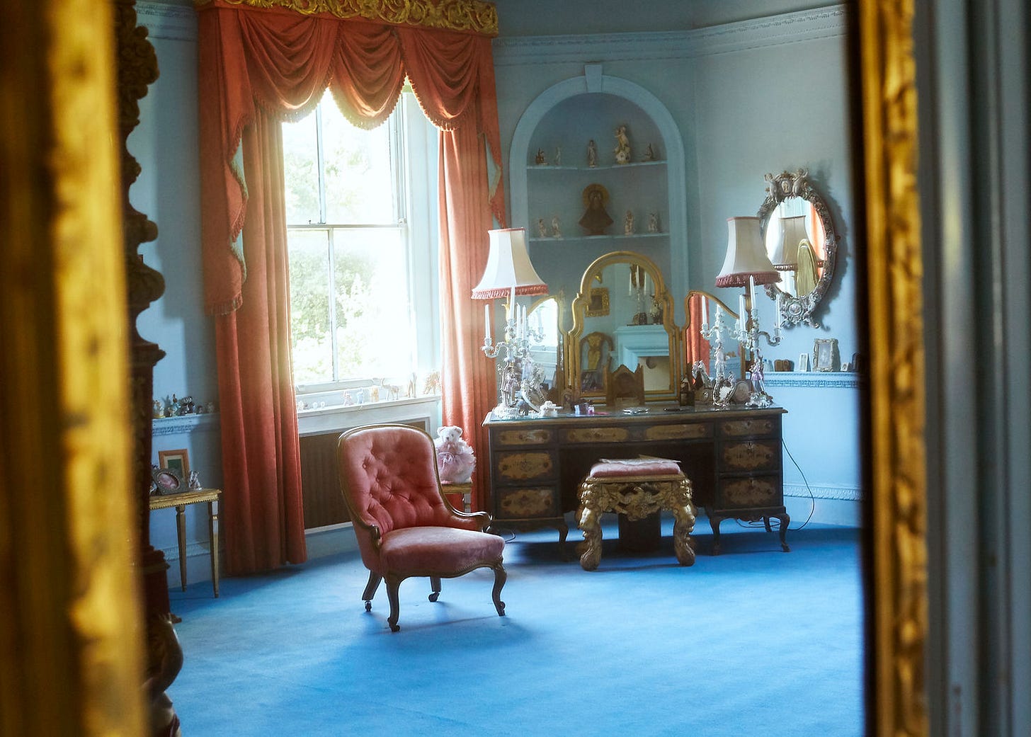 Elegant room with a pink chair, ornate dresser, and large mirror. Red curtains frame a window. Blue carpet and soft, vintage ambience. Dame Barbra Cartland's Bedroom Photographed By Claudia Legge