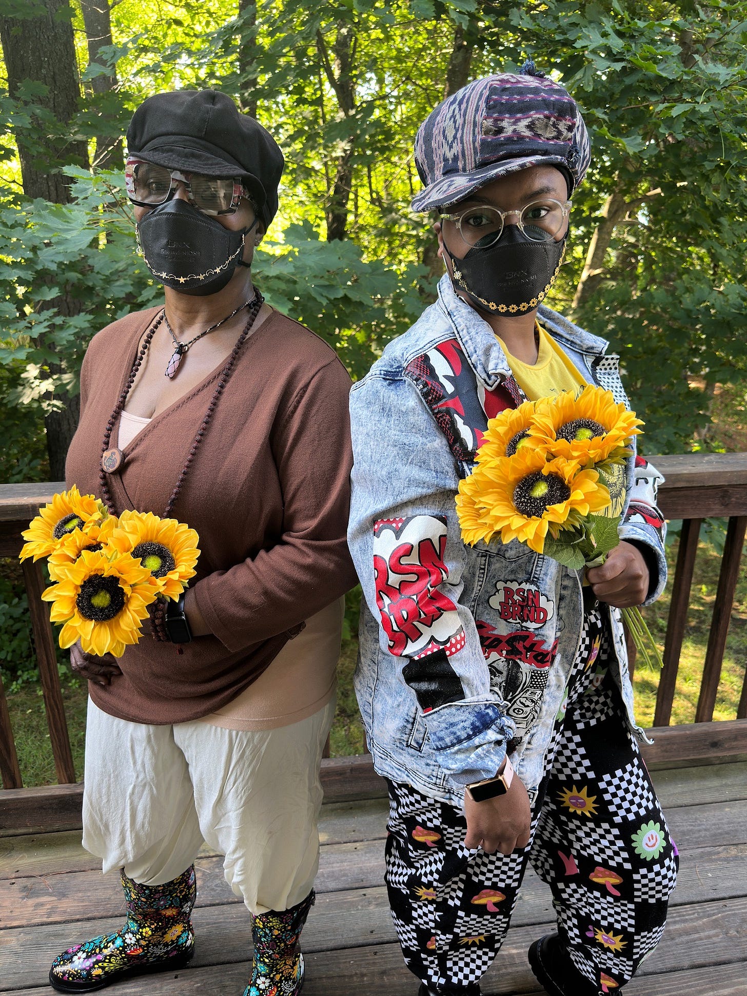 (ID: Amaranthia (young Black woman on the right) and Claire (older Black woman on the left both stand back-to-back. Both wear newsboy style caps, glasses and black N95 masks with mask chains. Both are on their back porch, holding yellow sunflowers. Amaranthia is wearing a blue denim jacket with comic style prints on it. Underneath is a yellow sunflower shirt. She wears black pants with checkered black and white patterns. Claire has a brown cardigan on with a white shirt under it and white pants. Behind them are some trees with lots of greenery. (ID: Amaranthia (young Black woman on the right) and Claire (older Black woman on the left both stand back-to-back. Both wear newsboy style caps, glasses and black N95 masks with mask chains. Both are on their back porch, holding yellow sunflowers. Amaranthia is wearing a blue denim jacket with comic style prints on it. Underneath is a yellow sunflower shirt. She wears black pants with checkered black and white patterns. Claire has a brown cardigan on with a white shirt under it and white pants. Behind them are some trees with lots of greenery.