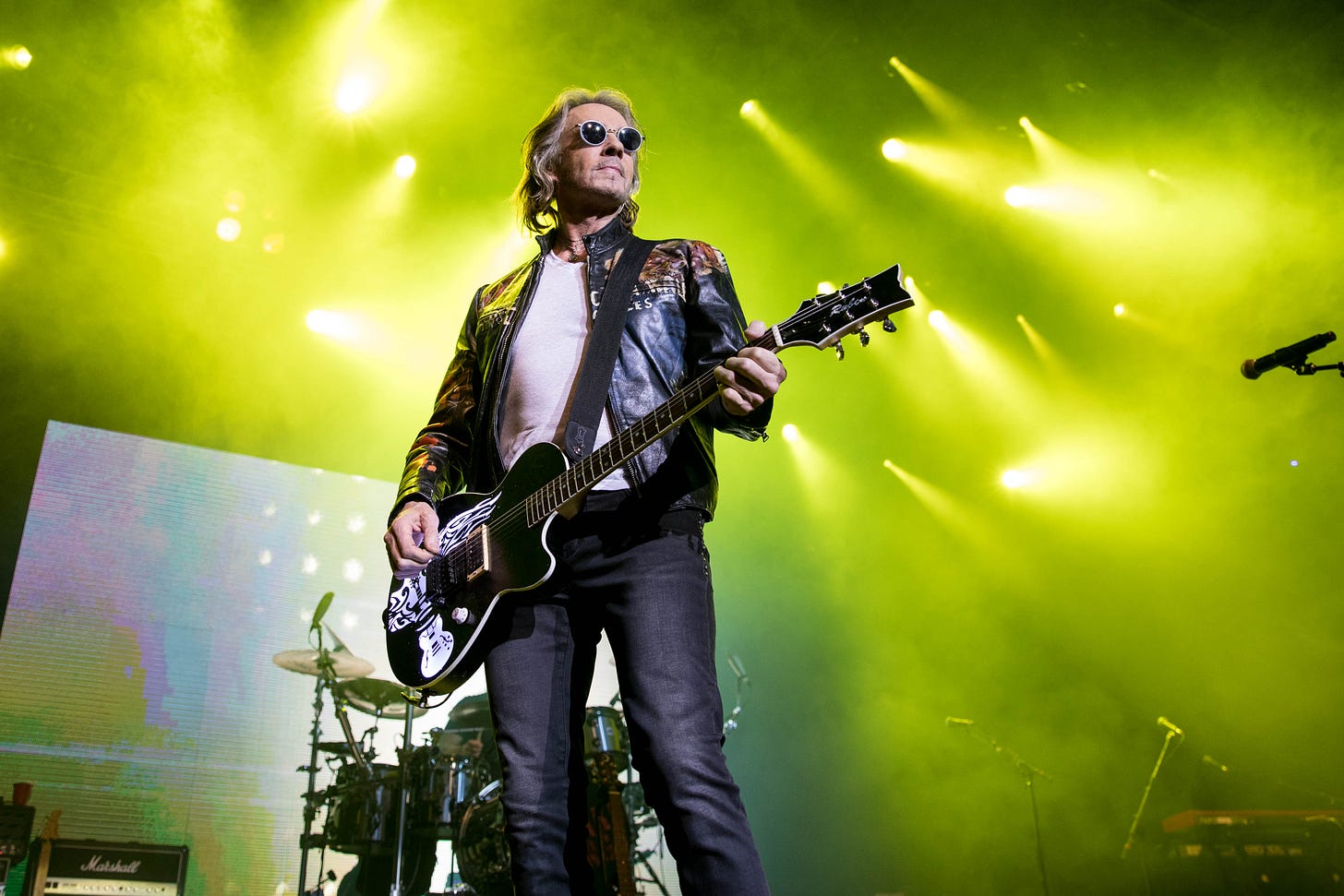 Rick Springfield performs on stage under bright yellow stage lights, wearing sunglasses, a leather jacket, and holding a black electric guitar with a custom graphic. A drum kit and amps are visible in the background.