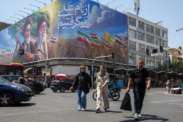 A large mural on a building shows two bearded men and a crowd with flags. Below, three people walk across a busy street with cars and traffic lights. A large mural on a building shows two bearded men and a crowd with flags. Below, three people walk across a busy street with cars and traffic lights.