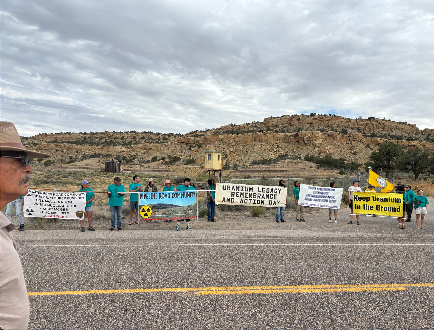 a small crowd lines up banners on the side of a street in the desert that read “uranium legacy day” and “keep uranium in the ground"