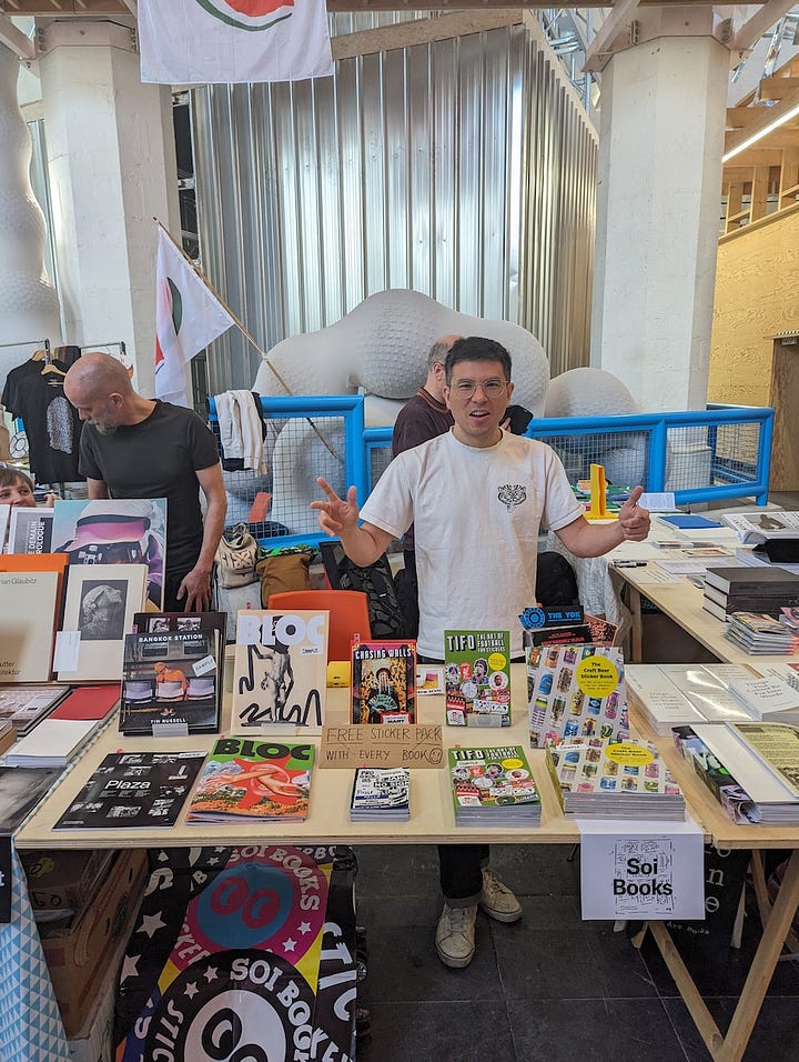 The first image is a poster for the Ghent Art Book Fair, held at Kunsthal Gent on May 11-12, 2024. The poster features bold black text with abstract design elements and lists various participating exhibitors and collaborators, such as Athena! Clay Fair, Imposed Alphabet, and Kero Zine. The second image shows an indoor art book fair scene with high ceilings and colorful tapestries on the walls, featuring multiple booths and tables where people browse through a variety of art books. The space has a grand, church-like architectural style with large windows, wooden beams, and hanging sculptures. In the third image, a person stands behind a booth at the Ghent Art Book Fair, displaying several art books and sticker packs, with a sign indicating that a free sticker pack comes with every book. The table includes works from Soi Books, with a variety of zines, books, and promotional items laid out. The fourth image captures two booth operators from the publishing house Grafis Nusantara sitting behind a table that displays various colorful books, zines, and art publications. One of them is making a peace sign while the other is smiling casually, creating a laid-back and friendly atmosphere at the event.