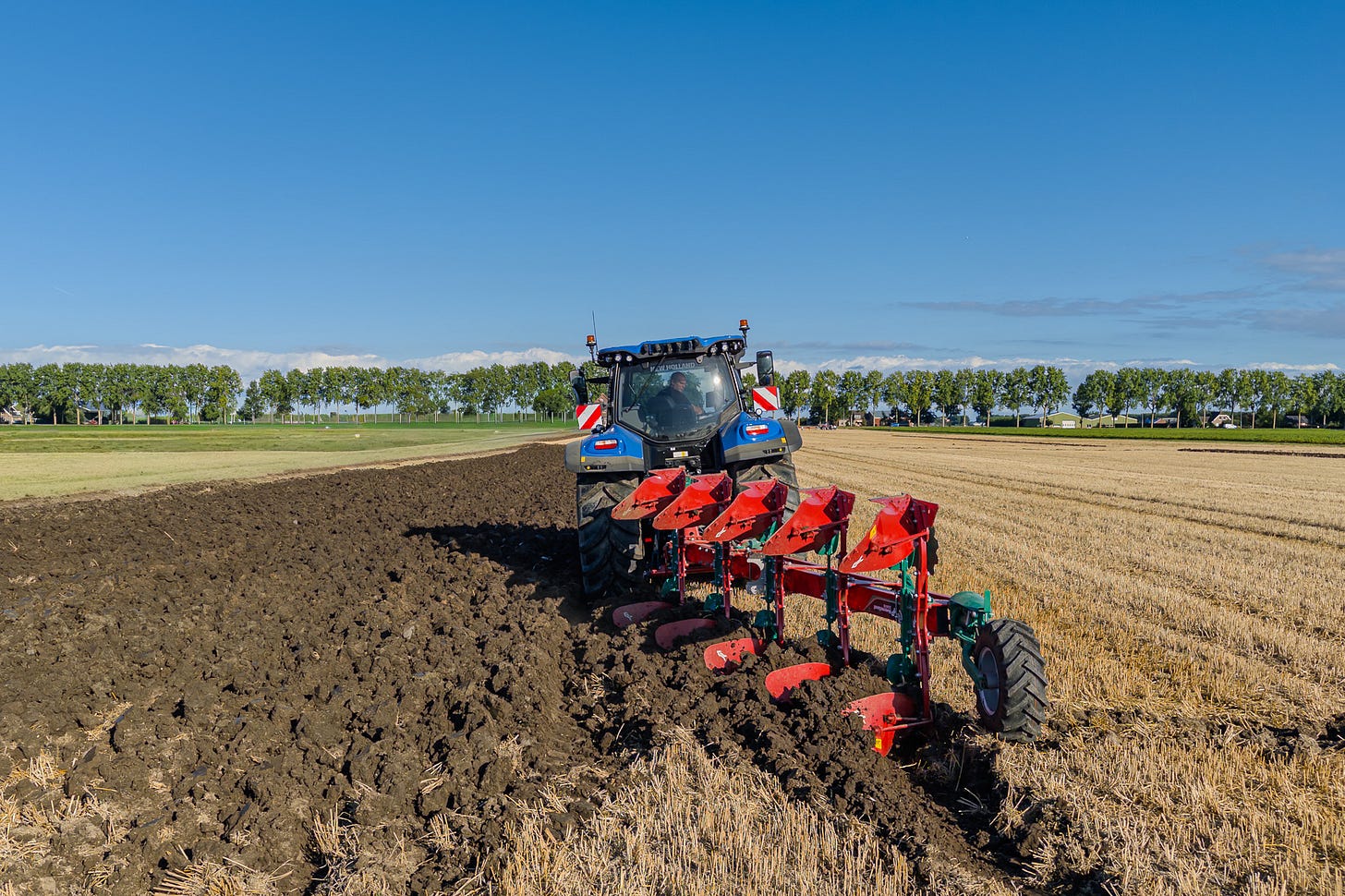 Ein moderner blauer Traktor von New Holland arbeitet auf einem abgeernteten Feld mit einem roten Anbaupflug. Die Maschine ist von hinten zu sehen, während der Pflug die obere Bodenschicht wendet: Links entsteht ein breiter Streifen frisch gepflügter, dunkler Erde, rechts bleibt das goldbraune Stoppelfeld stehen. Im Hintergrund begrenzt eine lange Baumreihe die offene Landschaft. Ein moderner blauer Traktor von New Holland arbeitet auf einem abgeernteten Feld mit einem roten Anbaupflug. Die Maschine ist von hinten zu sehen, während der Pflug die obere Bodenschicht wendet: Links entsteht ein breiter Streifen frisch gepflügter, dunkler Erde, rechts bleibt das goldbraune Stoppelfeld stehen. Im Hintergrund begrenzt eine lange Baumreihe die offene Landschaft.