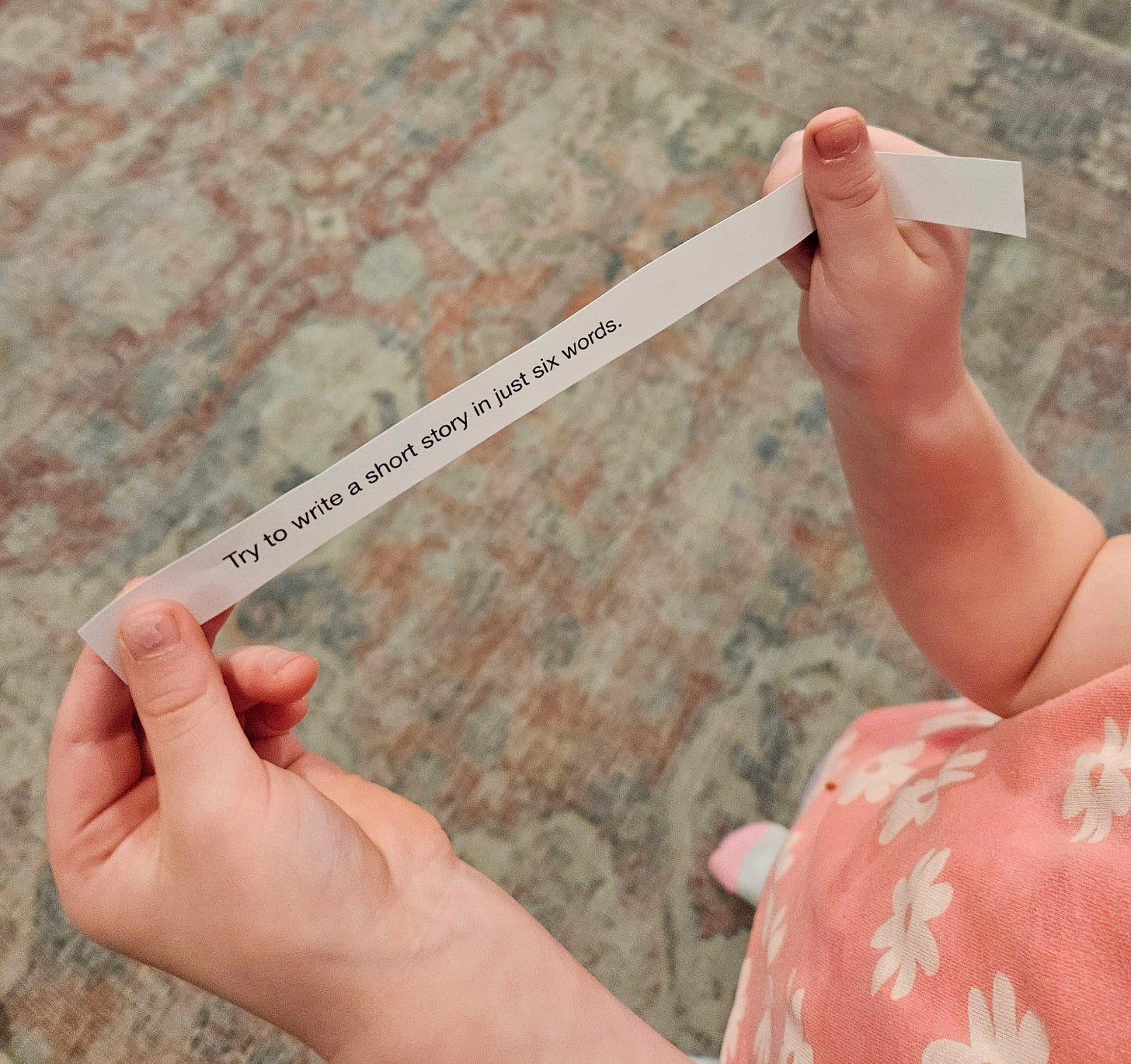 A young child and parent stretch open a Boredom Box slip together on the living room floor, revealing the prompt: 'Try to write a short story in just six words.