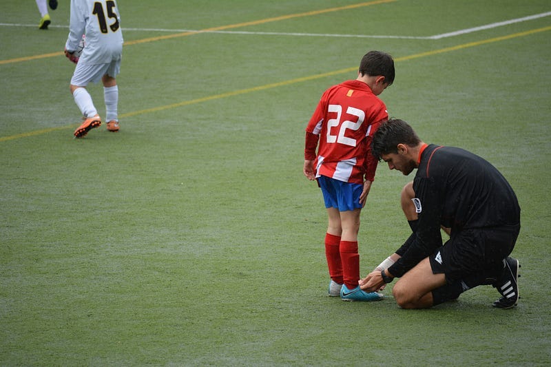 A man ties the shoe laces of a boy on the playing field.