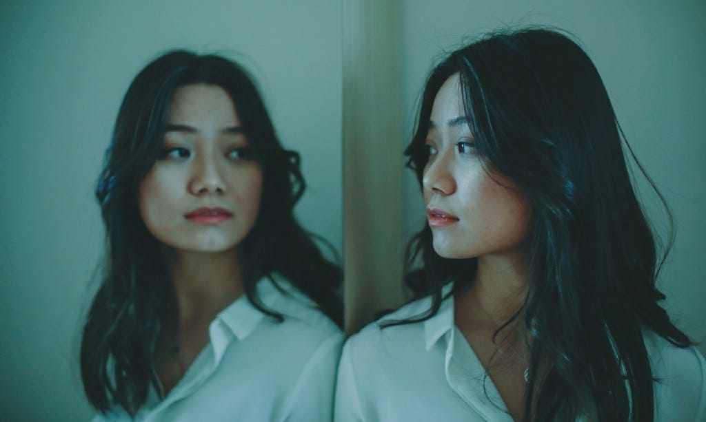 A woman with long black hair looking at herself in a mirror in dim lighting. A woman with long black hair looking at herself in a mirror in dim lighting.
