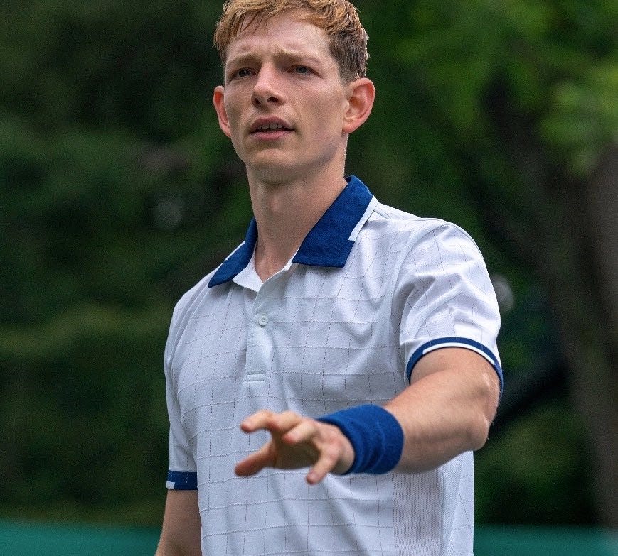 Mike Faist, depicted in a scene from the movie Challengers, with his arm outstretched in a white and bleu tennis uniform and matching blue wristbands.