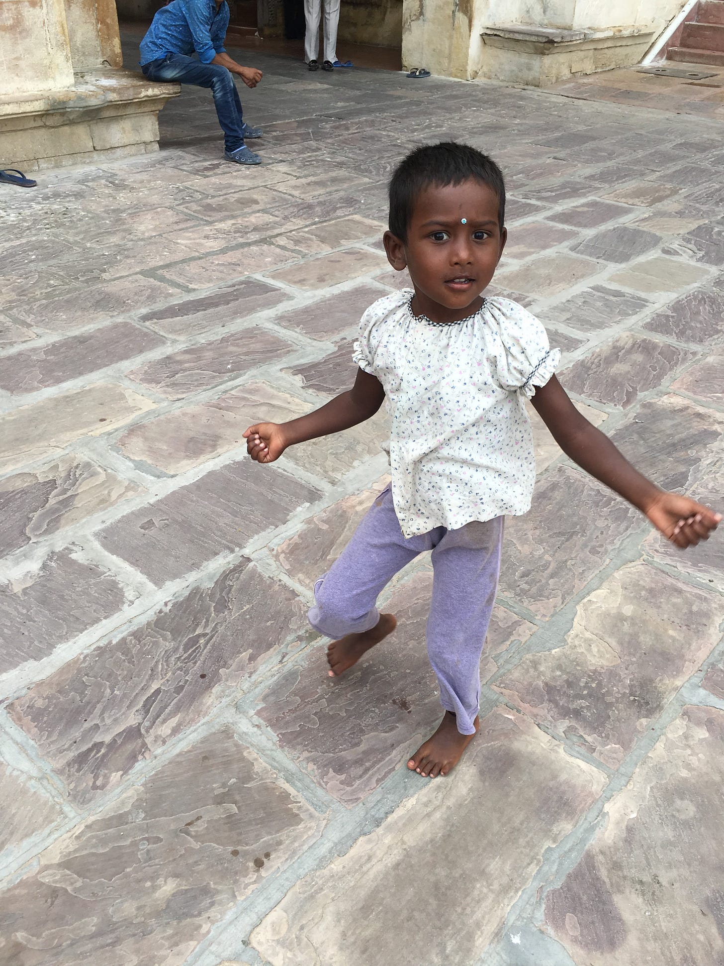 A three-year-old girl twirls in a stone and marble courtyard. She has very short black hair, dark brown skin, and dark eyes. She wears a white shirt and purple pants, looking off into the distance. A three-year-old girl twirls in a stone and marble courtyard. She has very short black hair, dark brown skin, and dark eyes. She wears a white shirt and purple pants, looking off into the distance.