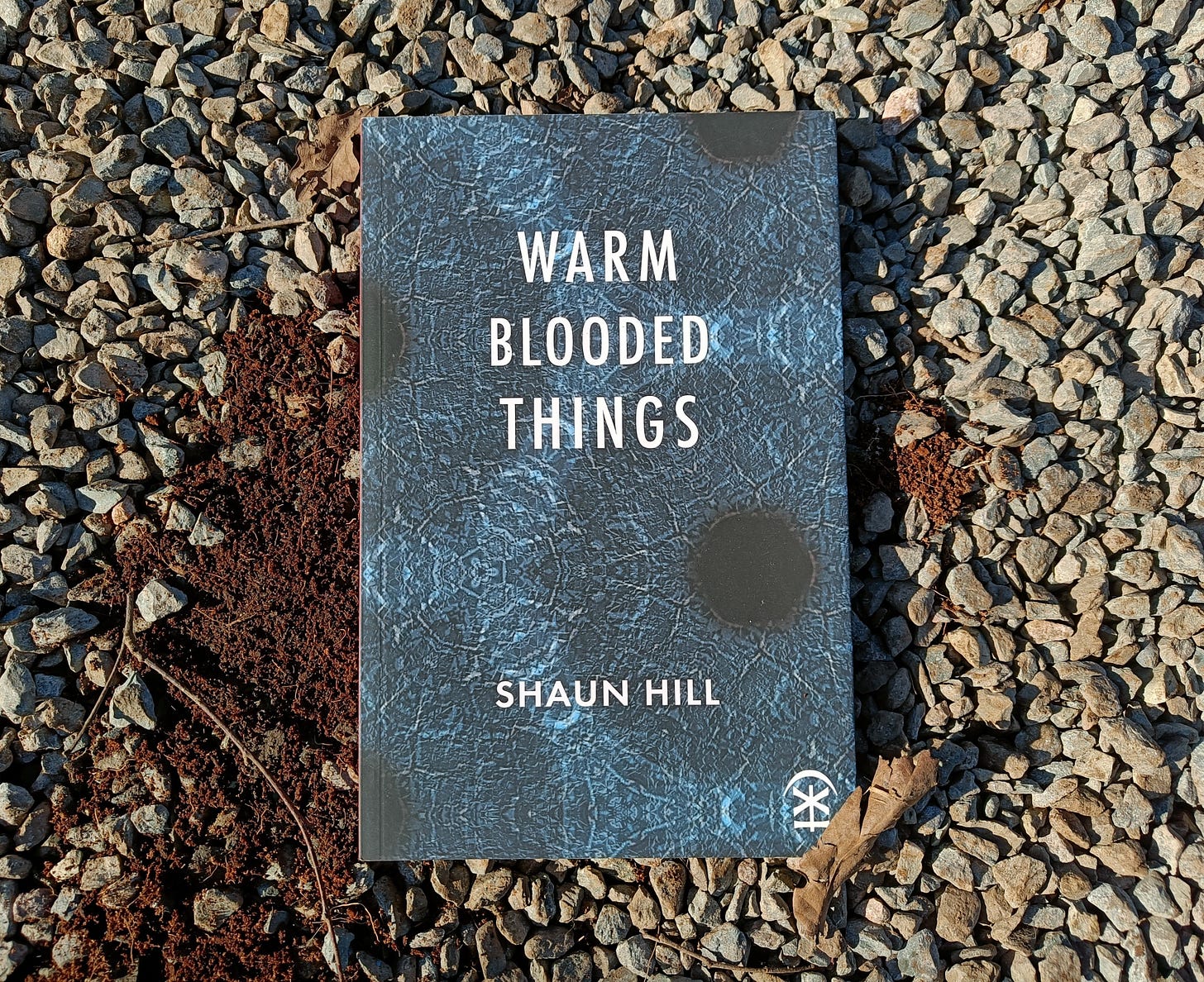 The poetry collection warm blooded things by Shaun Hill, published by Nine Arches Press, lying on grey gravel with soil visible to the left. The cover is dark teal with a weathered texture and circular shadow forms reminiscent of blood cells or tree rings. The title and author name in white capitals.