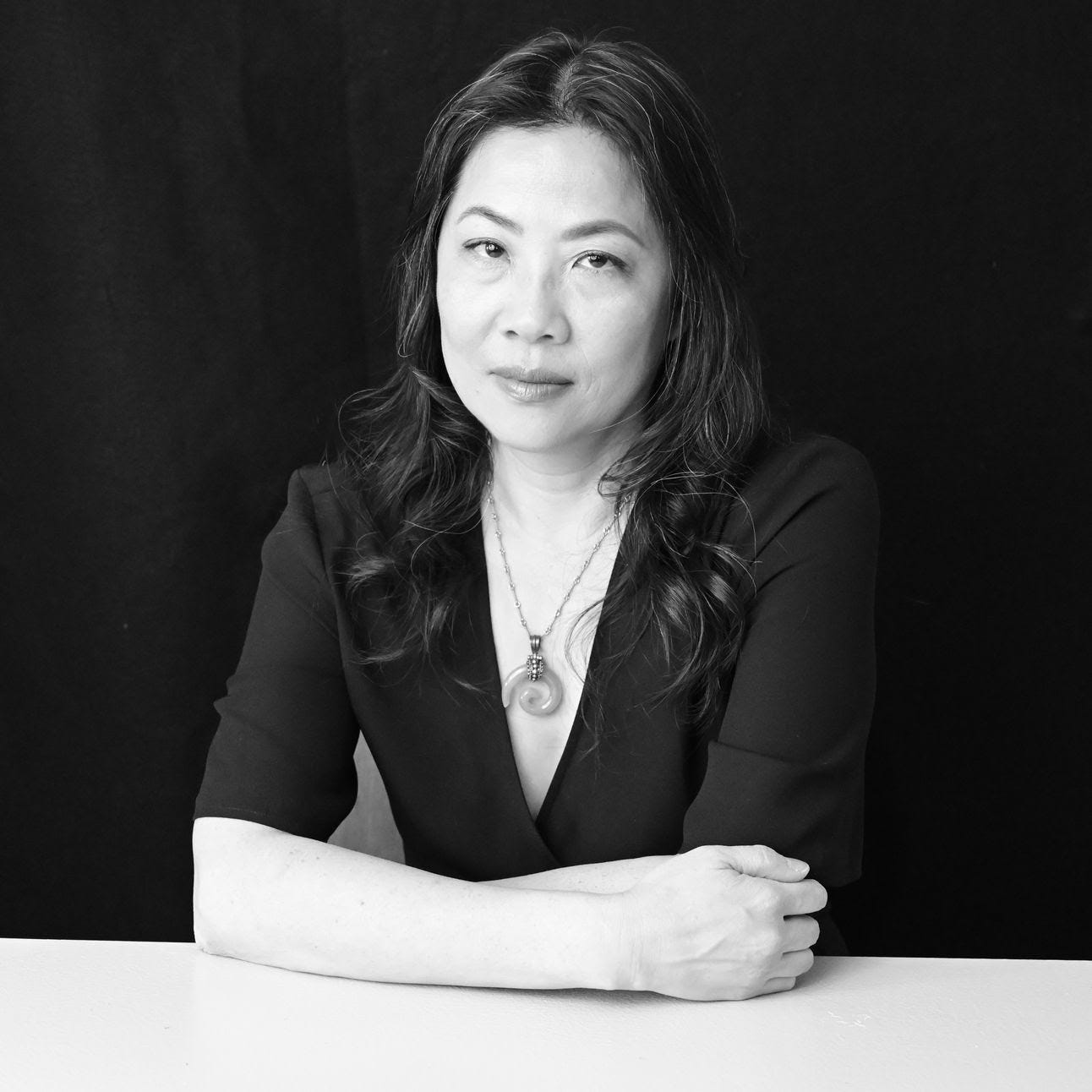 Black-and-white portrait of Karen Yin, a light-skinned Chinese American woman with long, wavy hair. Her arms are folded on a table in front of her. She smiles gently at the camera, with head tilted.