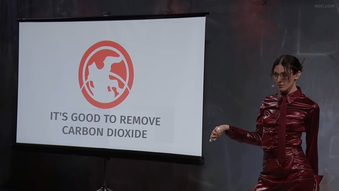A woman in a red leather outfit presenting on stage beside a slide reading "It's good to remove carbon dioxide".