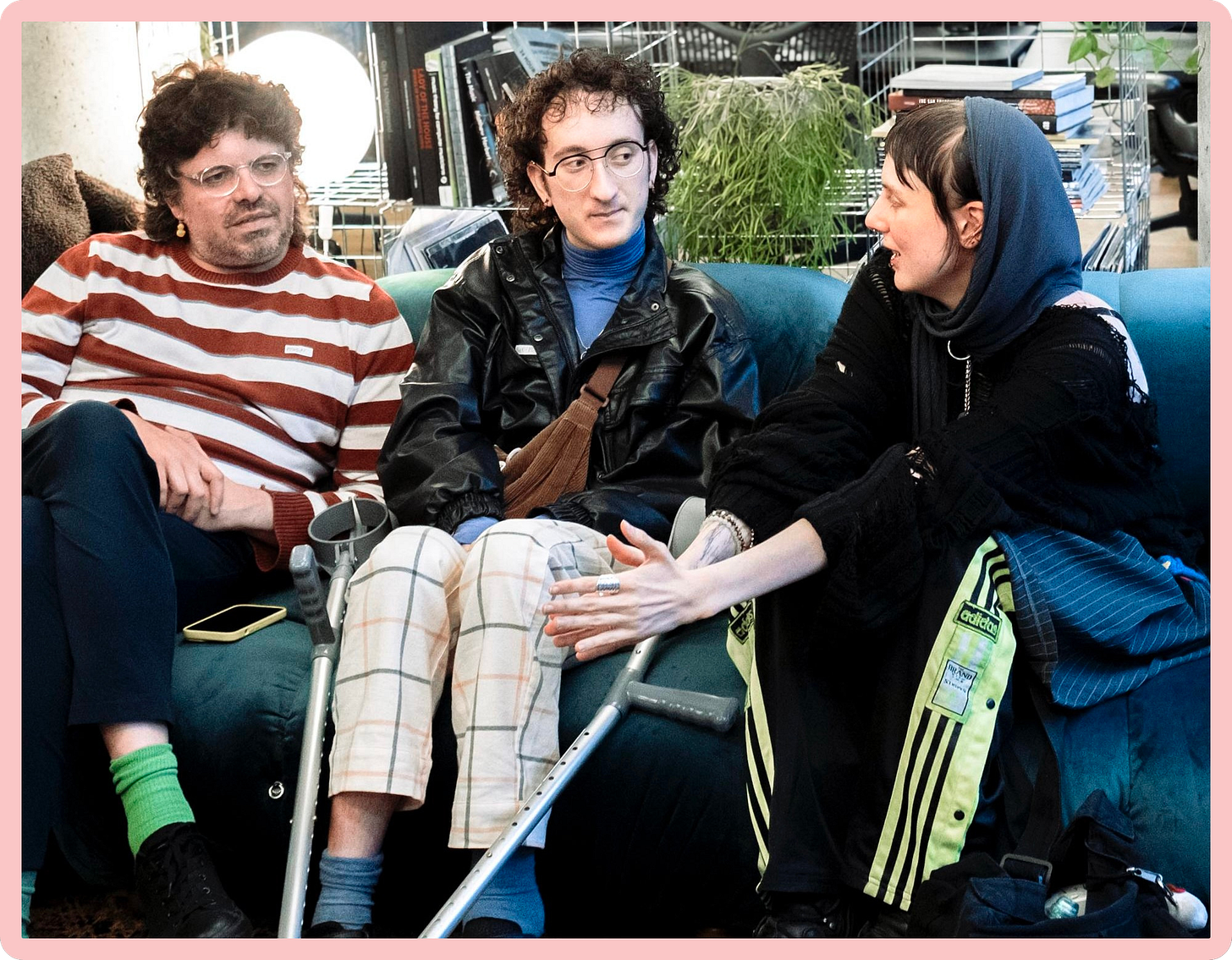 A photo of three young disabled people talking and sitting side-by-side on a blue sofa. Two of them are listening intently to a third person, who is talking. They are wearing colourful, bold outfits with interesting textures and patterns.