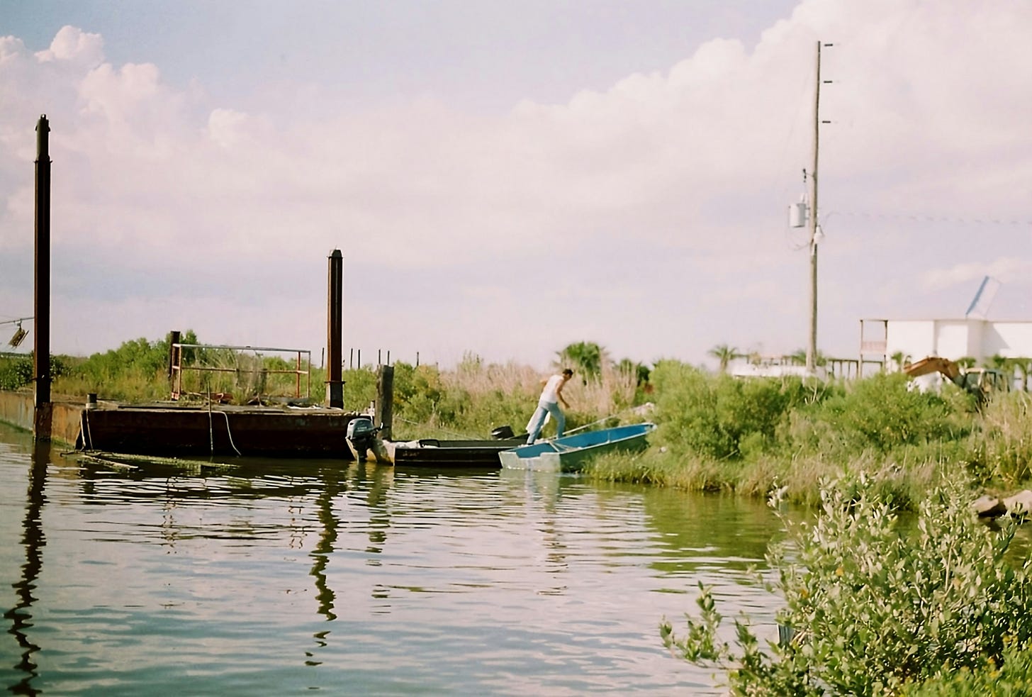 A lone figure guides a small boat toward the shallows beside a rusted barge in this tranquil bayou scene. Marsh grasses and soft, diffused sunlight evoke the rustic charm of rural wetlands, while the still water and open sky hint at a slow, unhurried pace of life. Captured on film, the image reveals a quiet resilience shaped by land and water, offering a cinematic glimpse into the heart of a place where nature and human endeavor converge.
