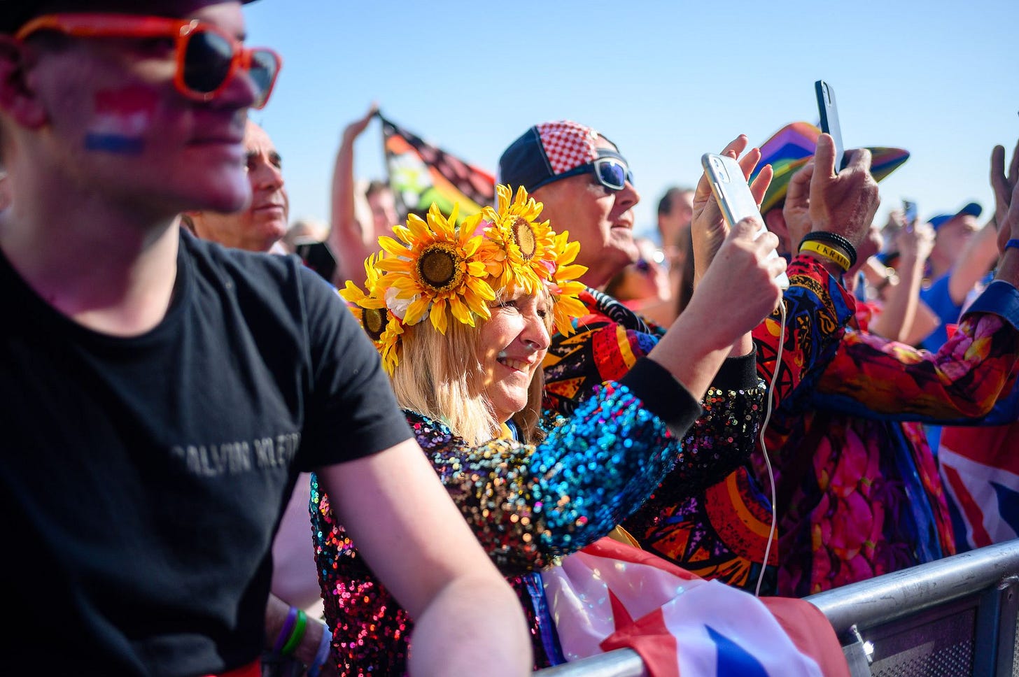 A woman in a rainbow sequin dress with a sunflower hat takes a photo in front of a stage.