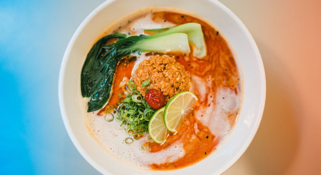 A plate of ramen with orange broth and leaves inside it, with an orange and blue background behind it