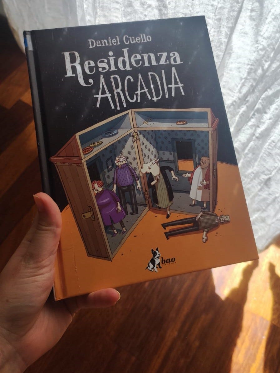 Una mano tiene un libro. Sullo sfondo parquet e una tenda bianca da cui entra un raggio di sole