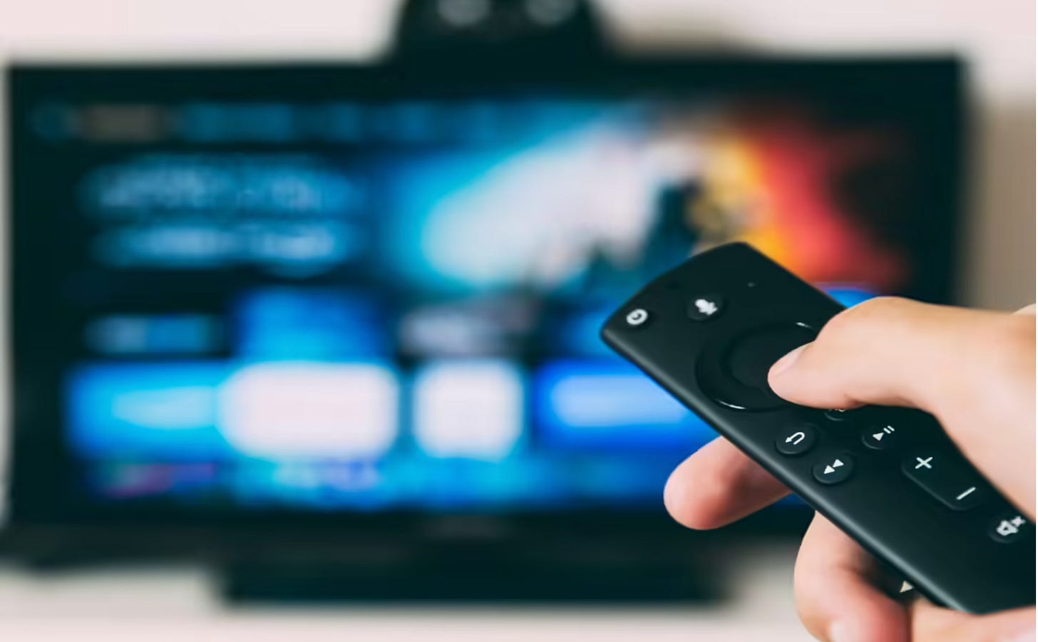 Close-up of a hand holding a TV remote, pointed at a blurred screen displaying vibrant colors. The scene conveys the action of choosing a program.