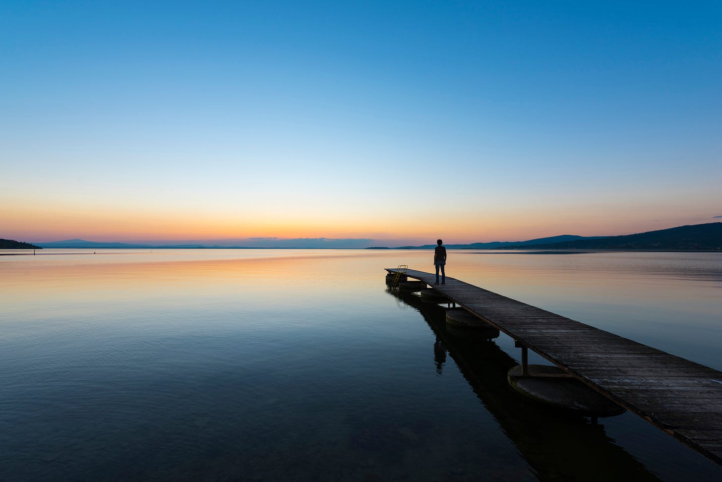 Lone figure on a short low pier at sunset over still water. Not the pier I pictured in the story but the solitude and color tones, slightly foreboding, got to me