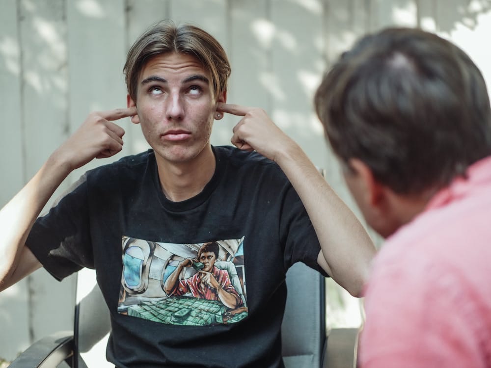 Free A teenage boy in a black shirt covers his ears and looks up, ignoring a conversation outdoors. Stock Photo