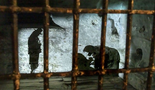 Two images showing cells at Colchester Castle. The first shows a wooden boarded room with cells on all sides. Cells have wooden doors and open barred windows. The second image shows the cell interior - wooden boarded stone rooms with silhouettes of prisoners projected on the wall.