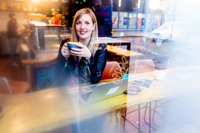 Lorraine is sitting in the window of a coffee shop holding a cup of coffee and smiling. She has long blonde hair and is smiling.