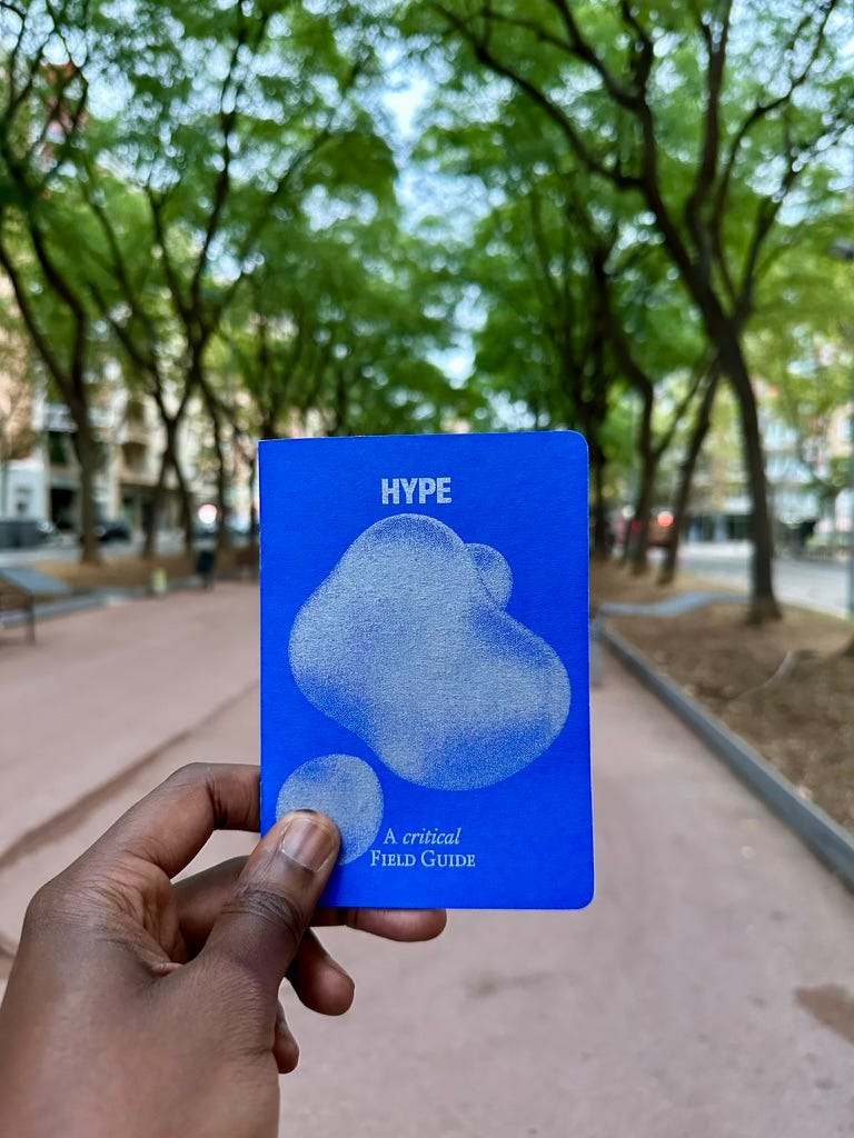 A Black manās hand holding a passport sized zine with a Yves Klein blue cover. The cover has a grey irregular bubble and the totle is HYPE: A Critical Field Guide. A Black manās hand holding a passport sized zine with a Yves Klein blue cover. The cover has a grey irregular bubble and the totle is HYPE: A Critical Field Guide.