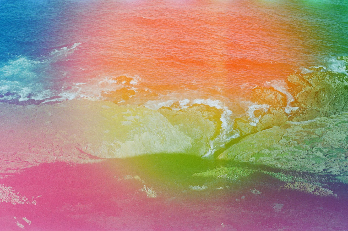 A film photo of the ocean and shore, with rainbow light leaks covering most of the frame.