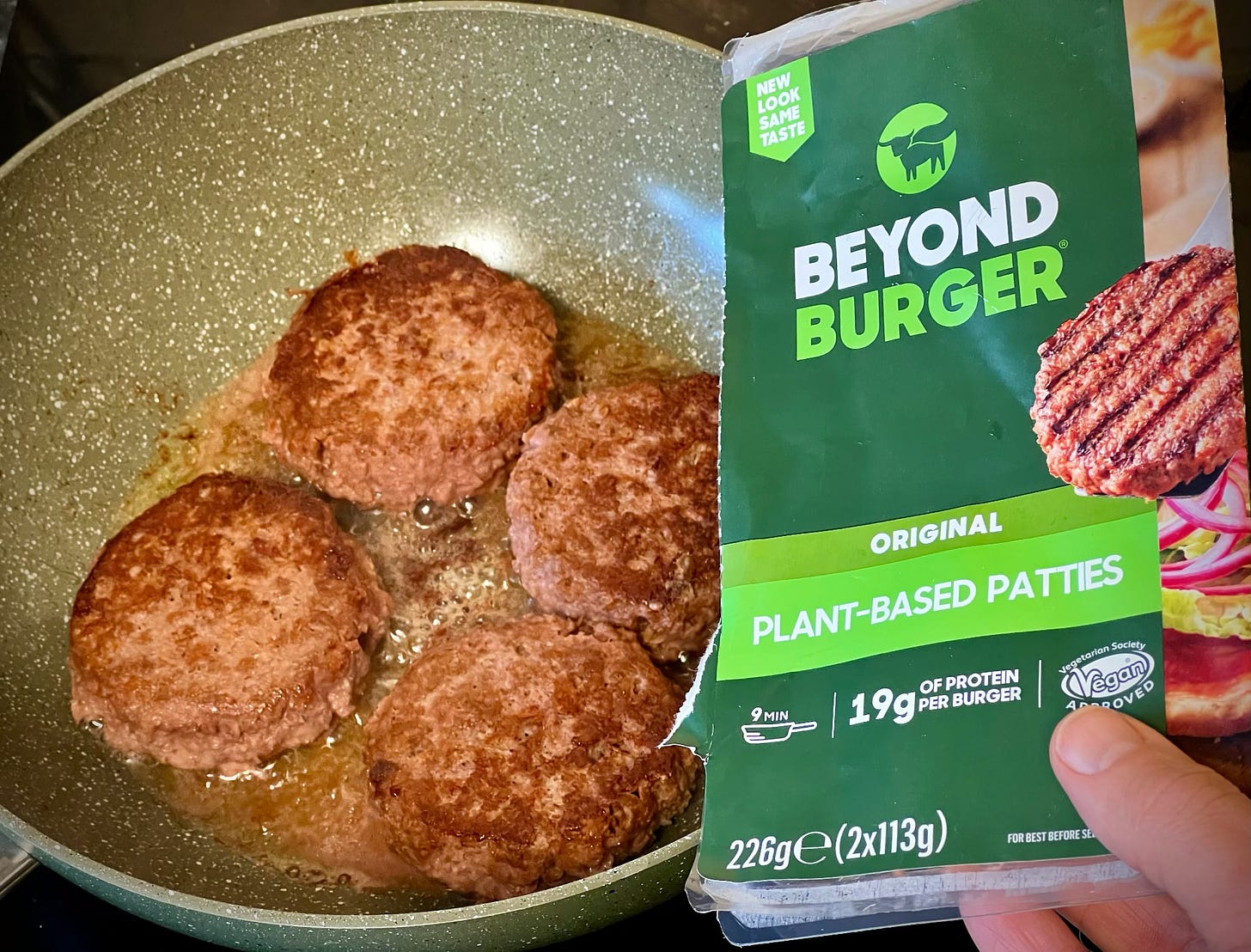 some sizzling fake burgers in a pan with the Beyond Burger packaging being held in hand.