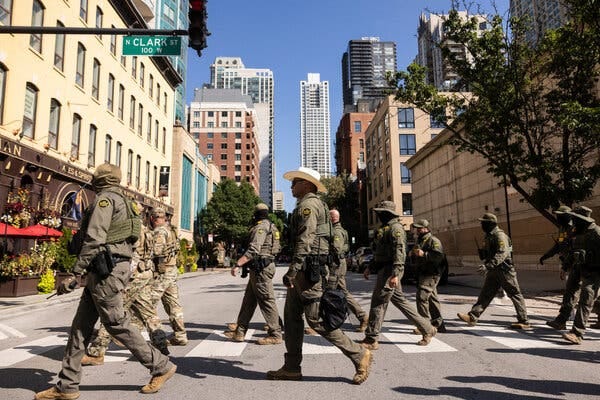 People in camouflage walk across a street near high-rise buildings.