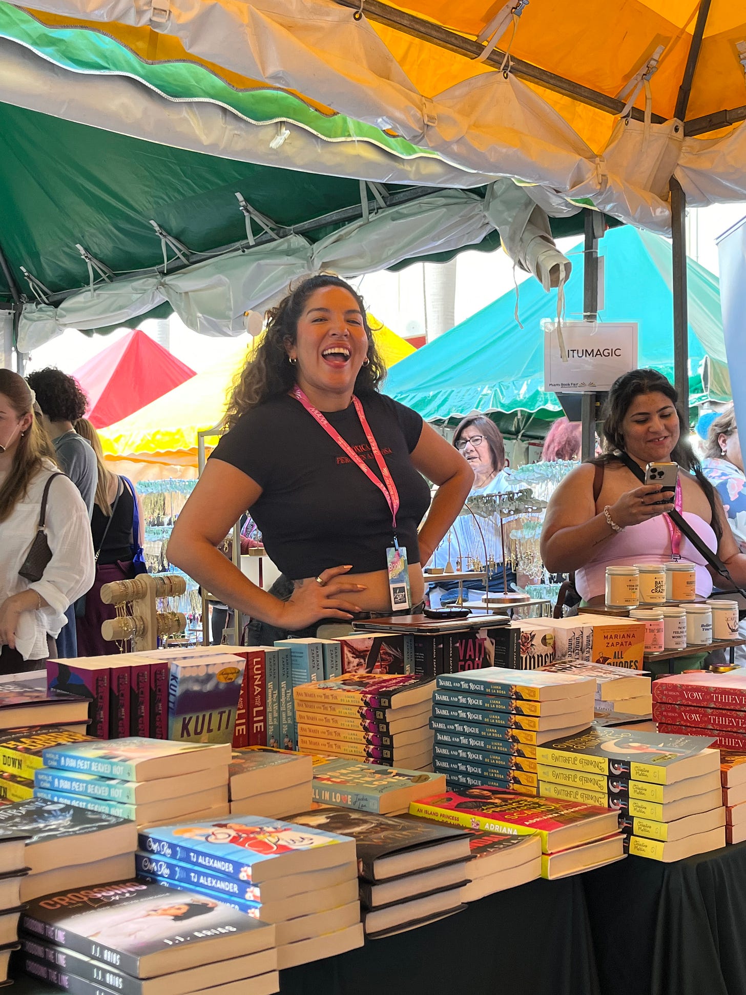 Photo of Latina woman with dark curly hair and a big smile with a jaunty hands-on-hips pose as she presides over a table of romance novels
