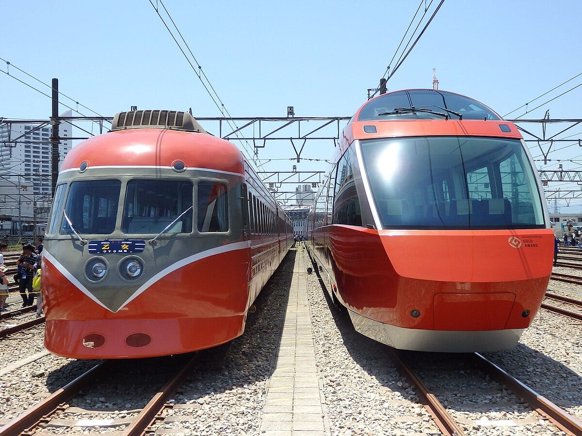 Side-by-side view of vintage and modern Odakyu Romancecar trains on parallel tracks under clear skies.