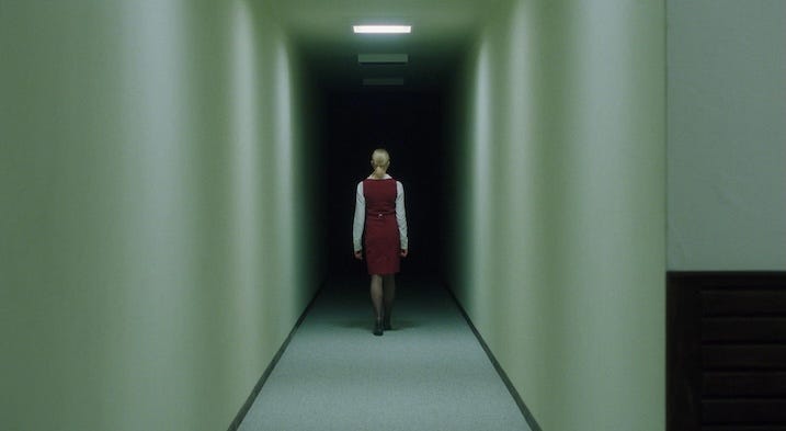 A woman in a red uniform walks down a stark, fluorescent-lit hallway that narrows into darkness. The symmetrical framing emphasizes her smallness and the oppressive atmosphere.