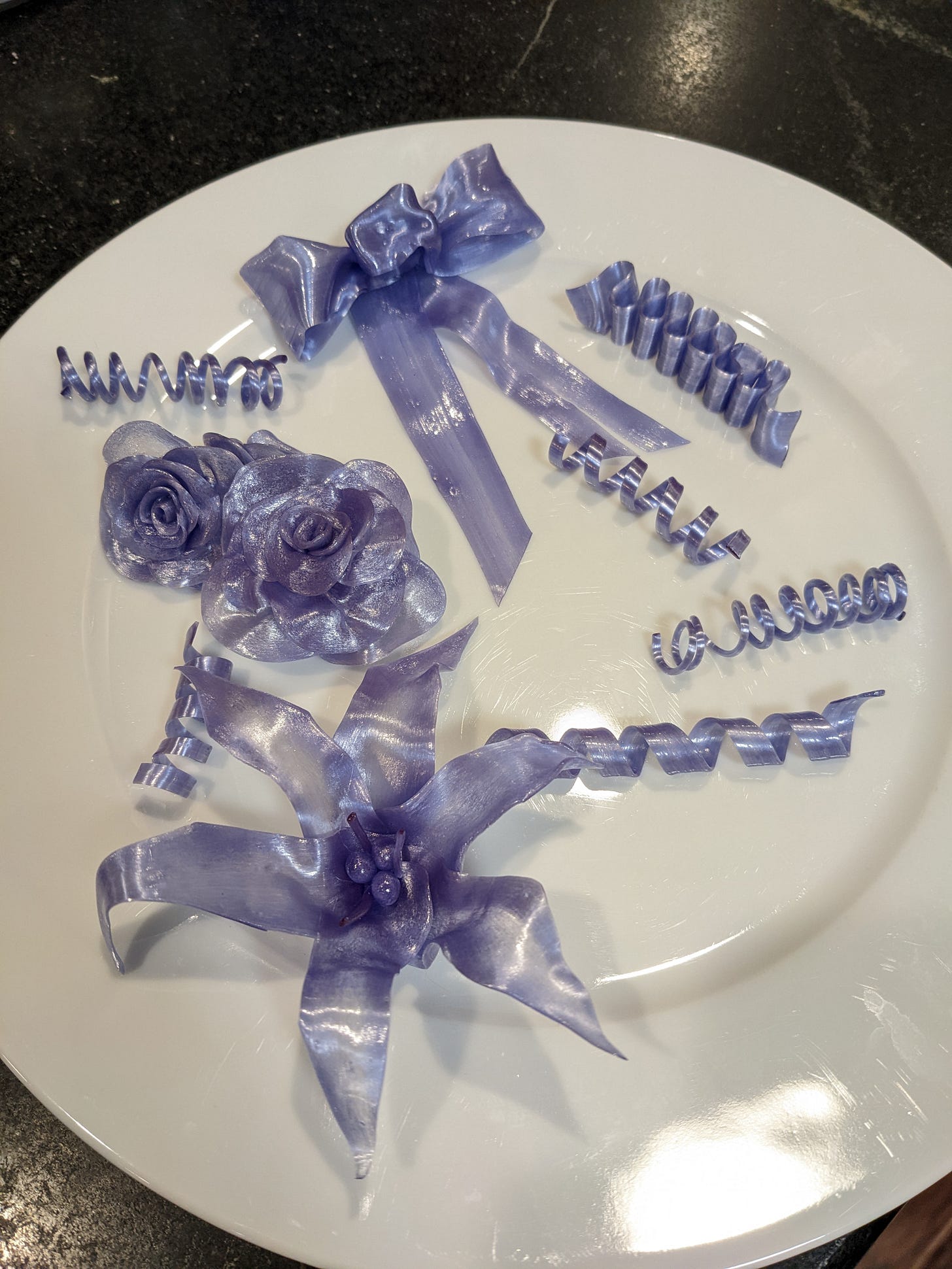 A white plate holding purple pulled sugar decorations, including ribbons, bows, roses, and a lily.