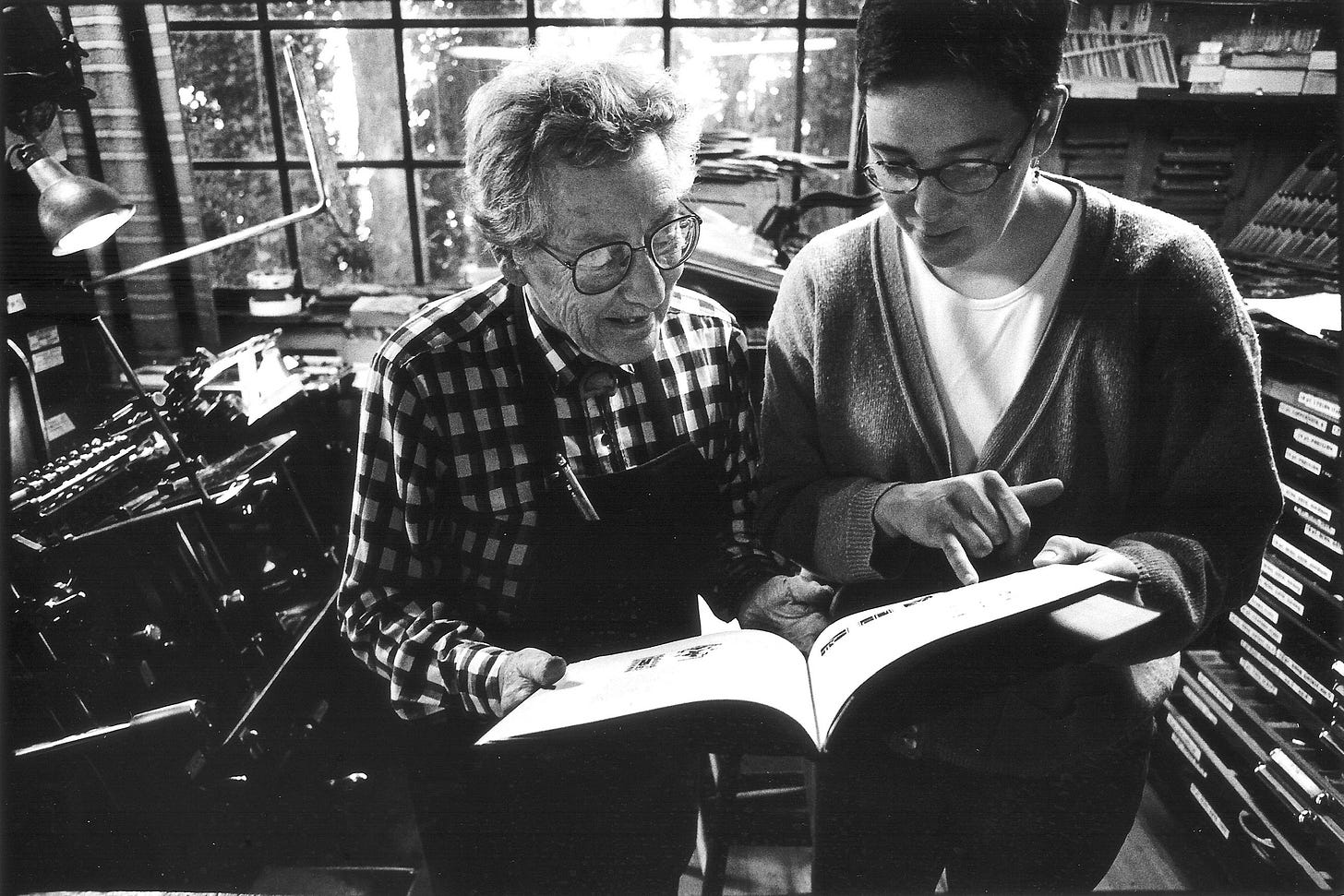 Black-and-white image of two women looking at a printed brochure, print shop in background