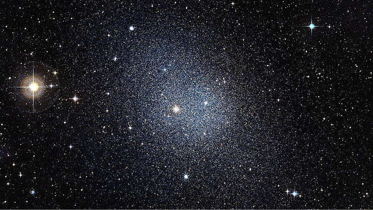 A dense star field with numerous stars against a dark background. The stars range from faint to bright, with a prominent cluster of stars forming a loose, spherical shape near the center. A bright yellow star is visible on the left side, and a bright blue star is on the right side. The central cluster is slightly more concentrated with a faint, hazy appearance, indicating a galaxy or star cluster. A dense star field with numerous stars against a dark background. The stars range from faint to bright, with a prominent cluster of stars forming a loose, spherical shape near the center. A bright yellow star is visible on the left side, and a bright blue star is on the right side. The central cluster is slightly more concentrated with a faint, hazy appearance, indicating a galaxy or star cluster.