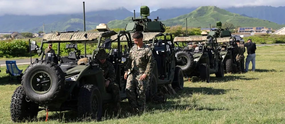 Military personnel in camouflage uniforms are working around a row of military utility vehicles (likely Polaris MRZR or similar light tactical vehicles) on a grassy field. The vehicles are equipped with various mounted systems including what appear to be sensor or weapons platforms on top. The setting appears to be a coastal or lowland area with green mountains visible in the background, consistent with Hawaii. Military personnel in camouflage uniforms are working around a row of military utility vehicles (likely Polaris MRZR or similar light tactical vehicles) on a grassy field. The vehicles are equipped with various mounted systems including what appear to be sensor or weapons platforms on top. The setting appears to be a coastal or lowland area with green mountains visible in the background, consistent with Hawaii.
