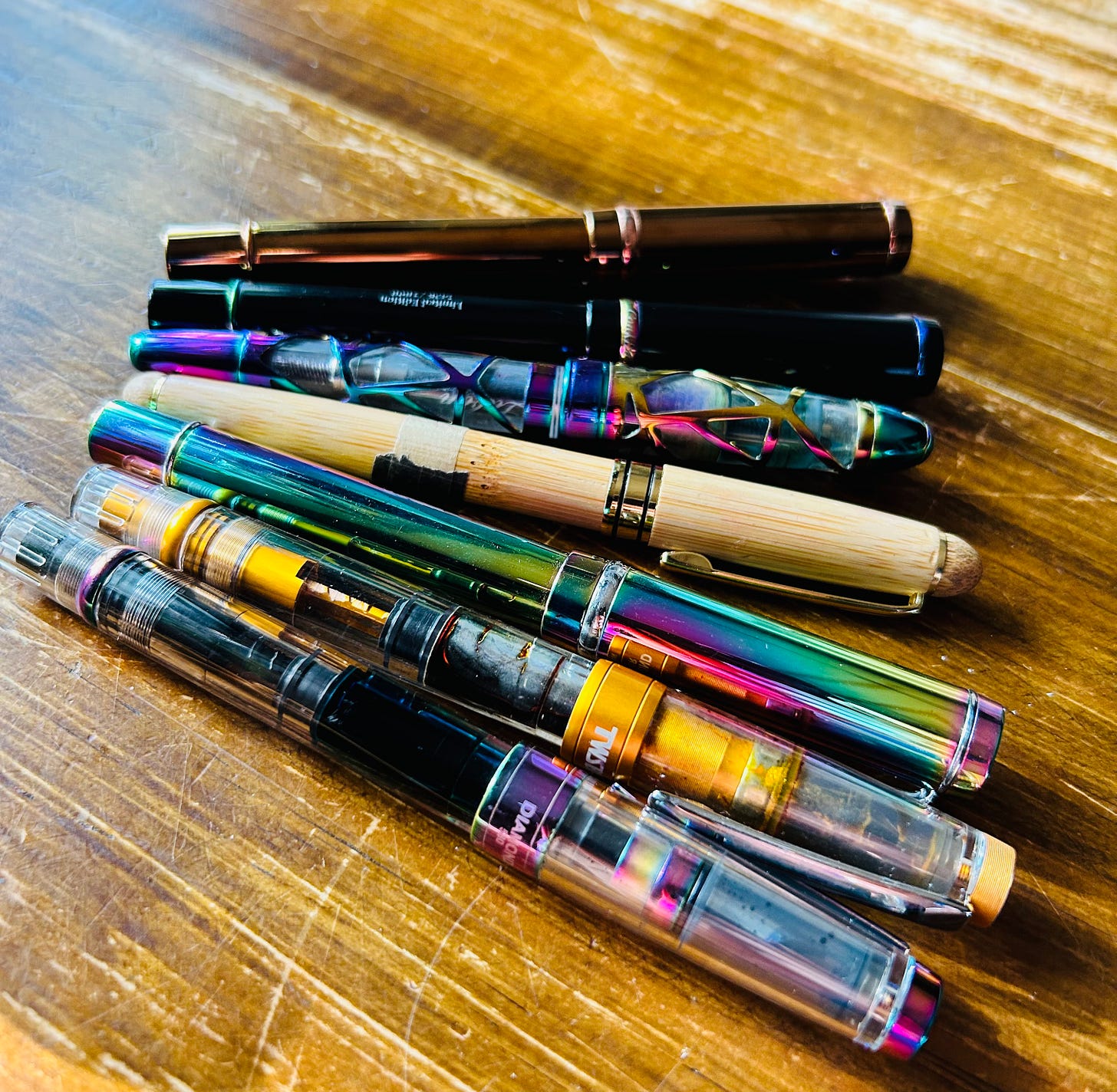 A collection of fountain pens laid on a wooden surface, including iridescent rainbow finish pens, clear demonstrator barrels, and a bamboo pen A collection of fountain pens laid on a wooden surface, including iridescent rainbow finish pens, clear demonstrator barrels, and a bamboo pen