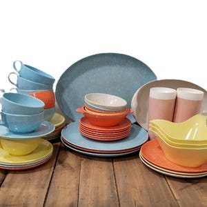May include: A collection of vintage dishware in various colors, including blue, orange, and yellow. The set includes cups, saucers, plates, bowls, and serving dishes. The dishware is arranged on a rustic wooden surface.