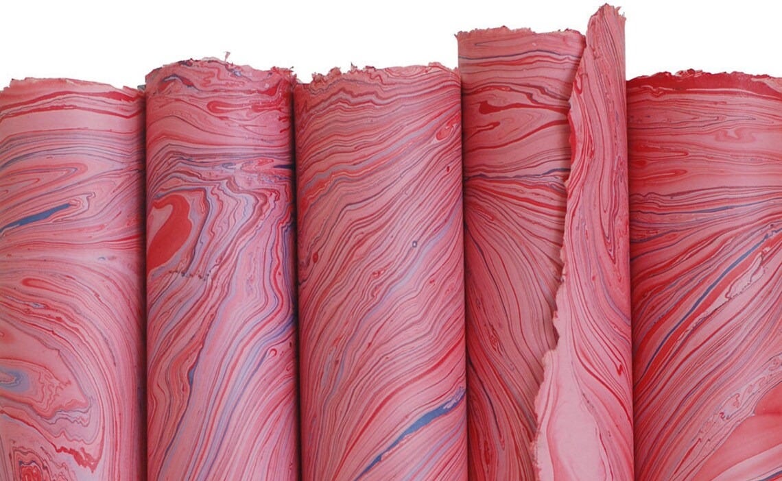 May include: Six rolls of marbled paper in shades of pink and blue. The paper is rolled and tied with a brown paper band that reads 'Sustain & Heal, 100% Fair Trade Paper for Planet Earth'.
