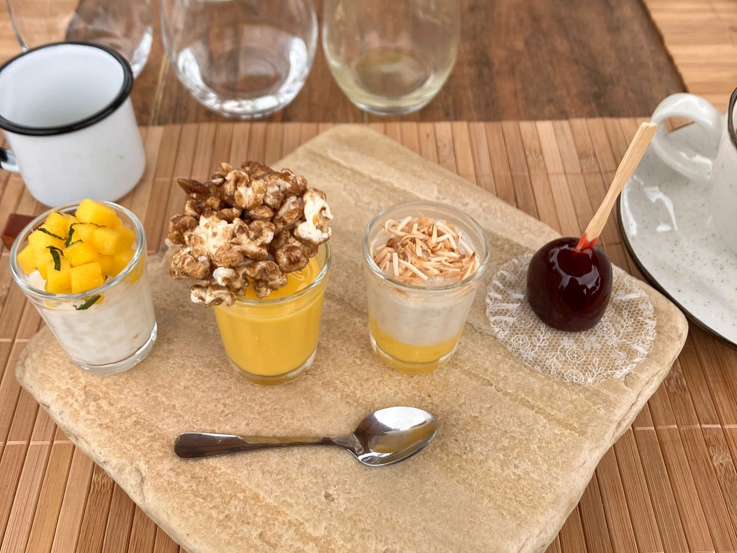 foto em close das sobremesas do brunch numa mesa de madeira. Enfileirados em mini copinhos de shot, da esquerda pra direita temos: sagu com creme de leite de castanha com pedacinhos de manga em cima com hortelã picadinha. curau com pipoca caramelizada em cima. arroz doce com geleia de mexerica. e na pontinha, um brigadeiro de vinho quente imitando uma maçã do amor. muito fofo.