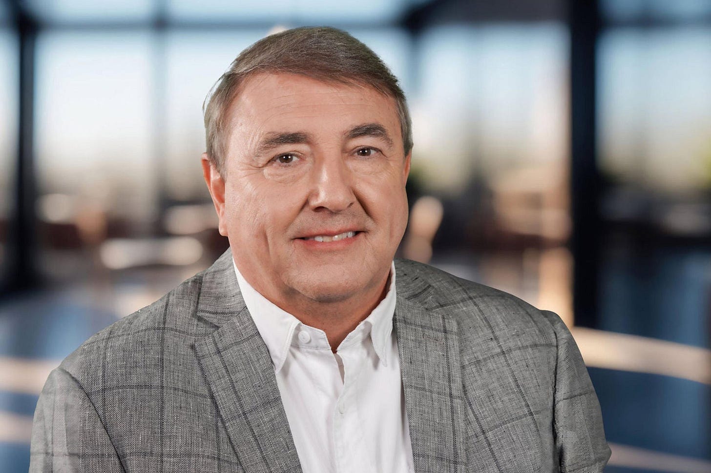 A man in a white collared shirt and gray patterned blazer smiles at the camera. The room behind him is in such soft focus that you can't make out any details. A man in a white collared shirt and gray patterned blazer smiles at the camera. The room behind him is in such soft focus that you can't make out any details.