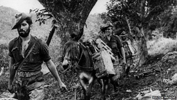 A black-and-white historical photograph showing three soldiers in wide-brimmed slouch hats trekking through a dense, muddy jungle trail. The soldier in the foreground has a thick beard and carries a rifle over his shoulder. Behind him, a mule heavily laden with wooden crates and supplies is led by another soldier. The environment is lush with large tropical trees, and the ground appears uneven and difficult to navigate.