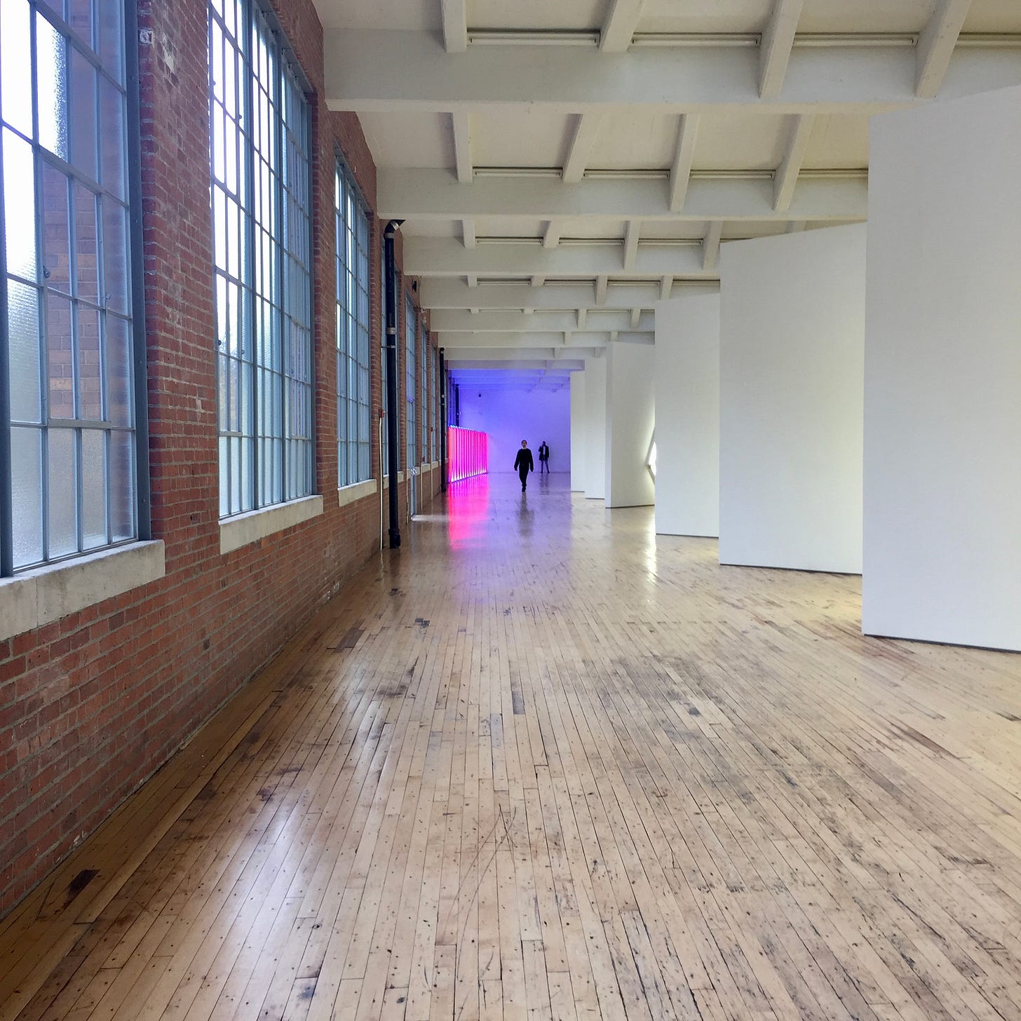 A long hallway with windows on the lefthand side, and two people standing at the end