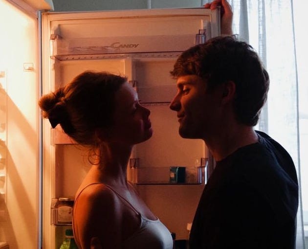 This may contain: a man and woman standing in front of an open refrigerator looking into each other's eyes