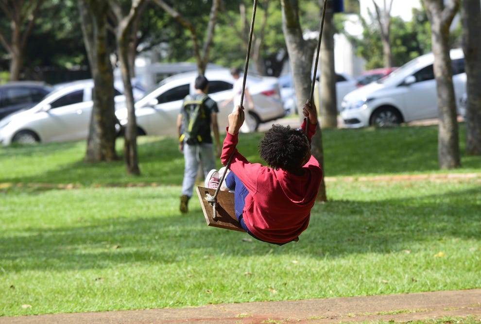 Criança de costas balançando em um balanço de madeira em um parque com gramado verde, árvores e veículos estacionados ao fundo, enquanto outra pessoa caminha em segundo plano.