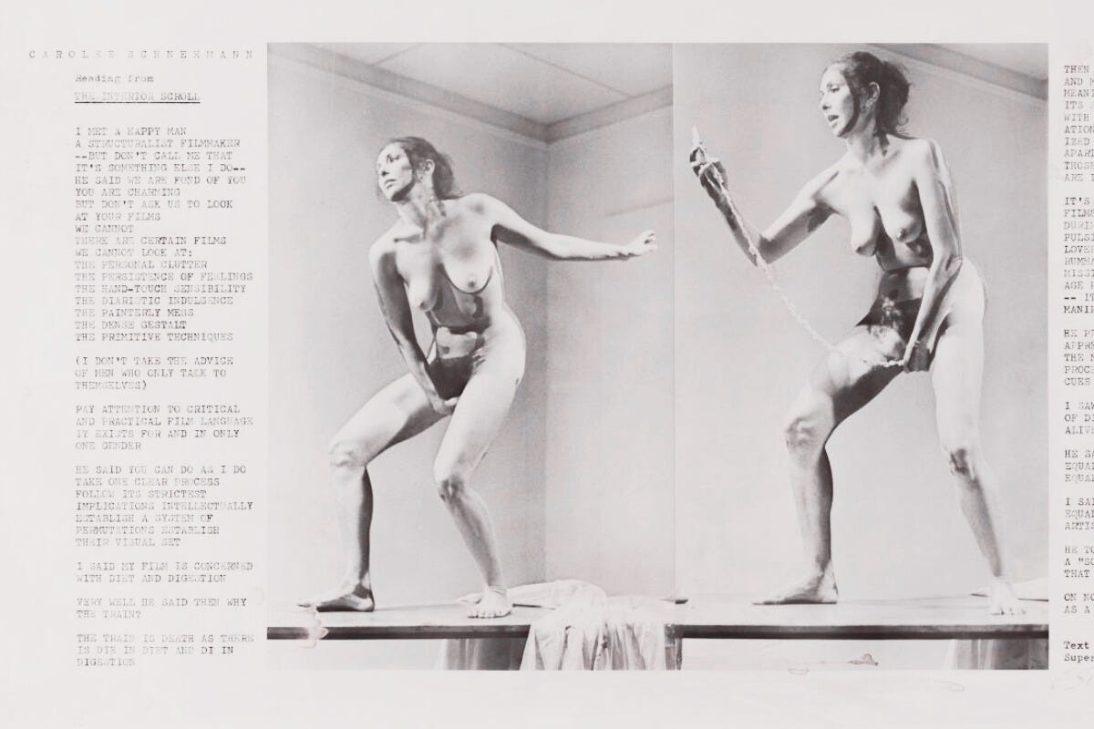Photograph of performance artist Carolee Schneemann standing nude on stage during her 1975 piece “Interior Scroll.” She holds a long paper scroll, partially unspooled from her vagina, while reading aloud from it.