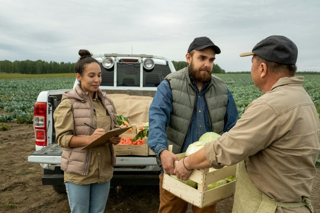 A man hands another man a box of produce in front of a pickup truck with the tailgate down. They are in a farm field and a woman with her hair in a bun looks on. Photo courtesy of Getty Images via Unsplash.