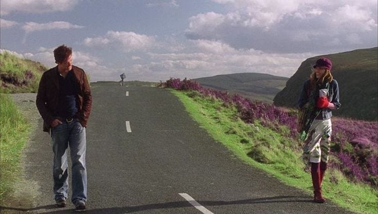 This may contain: two people standing on the side of a road with purple flowers in the grass behind them
