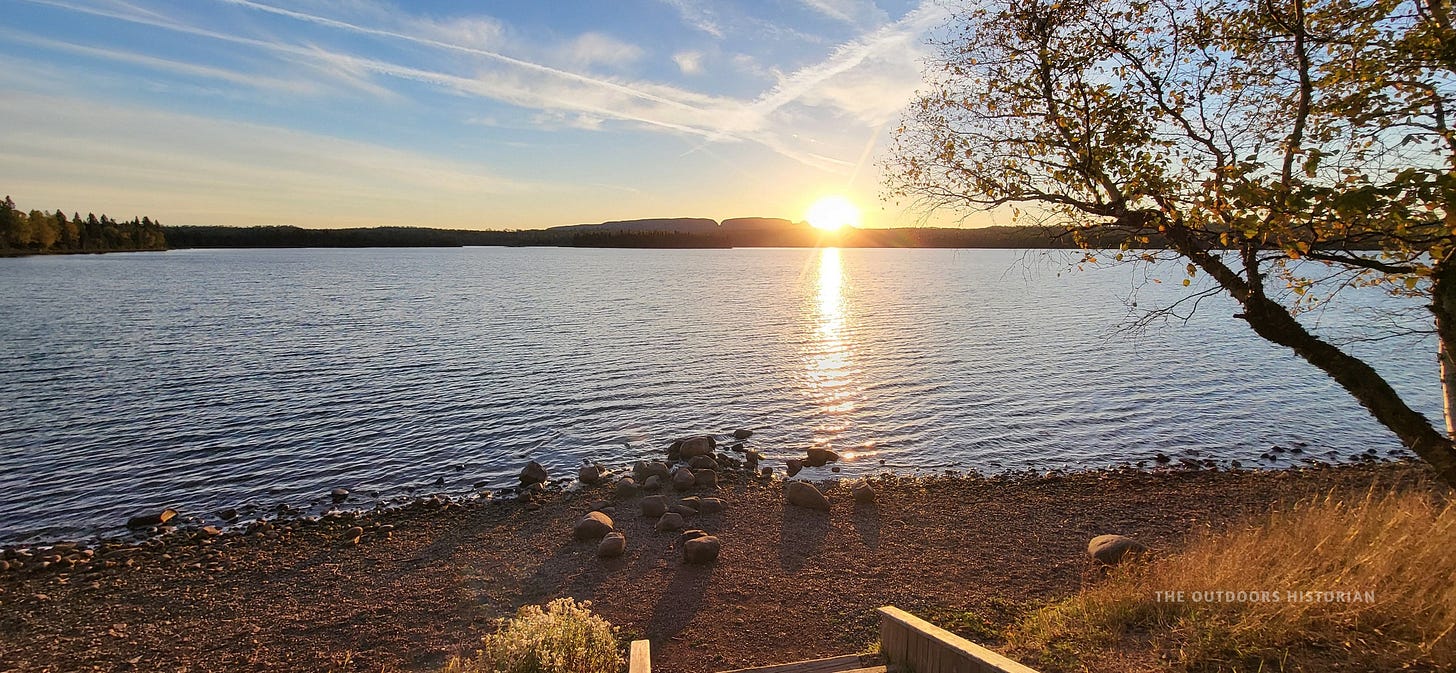 A peaceful sunset scene. The sky is streaked with orange and pink, reflecting on the calm water of a lake. The dark silhouette of the Sleeping Giant is in the distance.